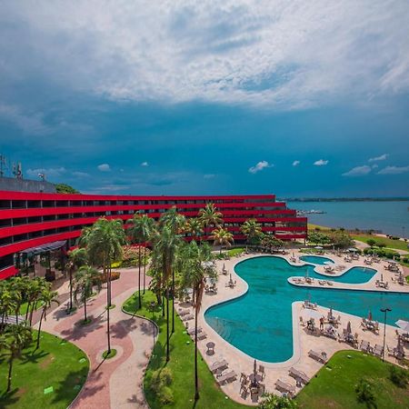 Отель Royal Tulip Brasilia Alvorada Экстерьер фото The photo shows a modern hotel building with a striking red exterior, surrounded by lush greenery. In the foreground, there is a large, creatively shaped swimming pool, with lounge chairs arranged around it. The backdrop features a body of water, pos